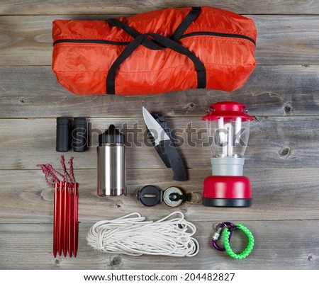 Overhead view of basic hiking gear placed on weathered wooden boards. Items include tent inside of bag, pegs, compass, canteen, rope, knife, case, lantern and binoculars.