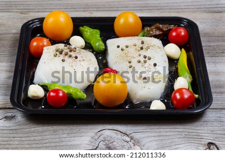Front view of fresh raw Cod Fish Fillets in cooking pan with tomatoes, garlic, peppers and peppercorn salt on rustic wooden boards