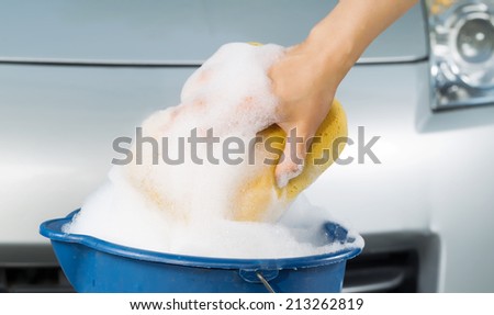Closeup of female hand holding soapy sponge out of the bucket with car in background