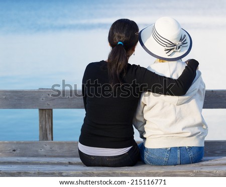 Closeup of mother and mature daughter siting on wooden bench while looking at the lake