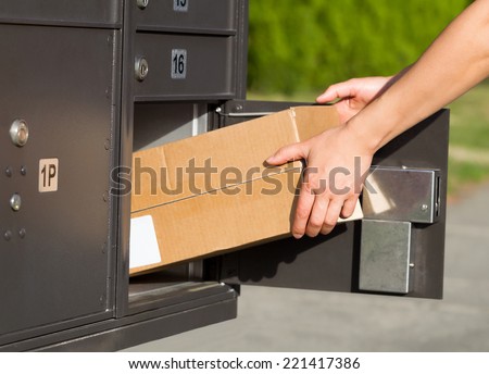 Horizontal image of female hands taking large package out of postal mailbox with green grass and sidewalk in background