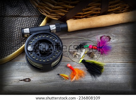 Close up top view of  fishing fly reel, landing net, creel and assorted flies, partial cork handled pole on rustic wooden boards with vignette border