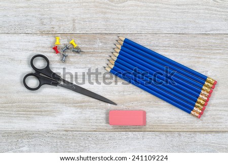 Top view of blue colored pencils, lined up, eraser, scissors, and pin tacks on wooden desktop