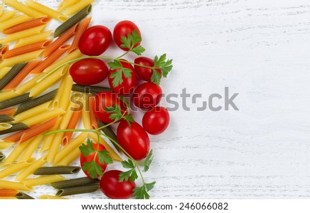 Raw pasta, of different colors, small grape tomatoes and parsley on rustic white wood. Top view angled shot.