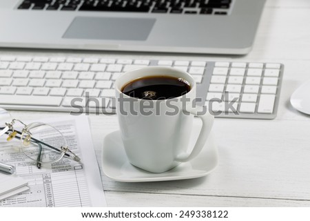 Close up of a fresh cup of black coffee with laptop, keyboard, pen, mouse, reading glasses and tax forms in background on white desk. Focus on front lip of coffee cup.