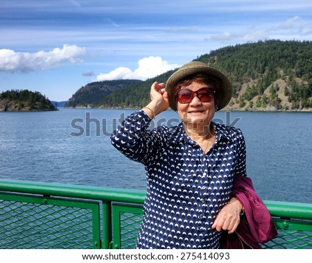 Senior woman, looking forward, wearing hat and sun glasses while standing on boat dock railing with ocean, mountain and blue sky in background