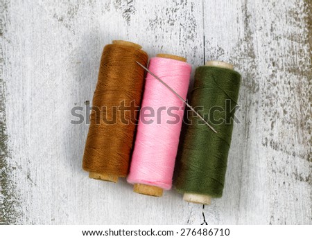 Three spools of different colors of thread, with single needle, on top of rustic white wood.