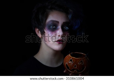Teen girl in scary makeup holding pumpkin container on black background. Trick or treat concept.