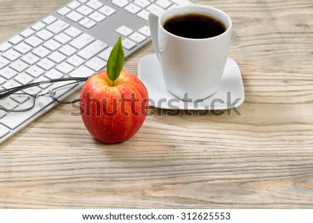 Close up of a red apple with computer keyboard, reading glasses and cup of coffee in background on desktop.