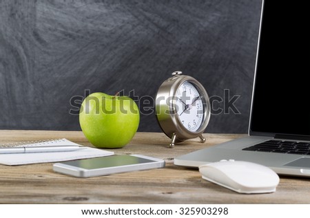 School desktop with laptop, mouse, clock, cell phone, notebook, pen and green apple in front of blackboard. Layout in horizontal format with copy space.