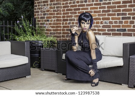 Sexy african american gothic woman smoking. Sitting on a couch outdoor patio, space for text.