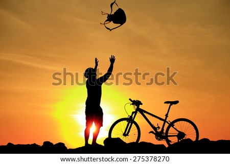 Silhouette of man and his bike in action throwing bag above his head on rock mountain with sunrise twilight background. Symbol of relax, success and touring.