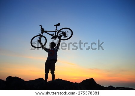 Silhouette of mighty man in action lifting bike above his head on rock mountain with sunrise twilight background. Symbol of relax, success and touring.