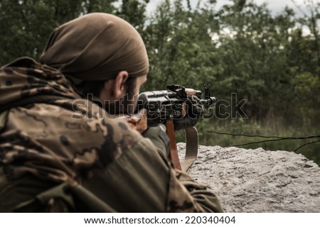 Rebel aiming an assault rifle at target, hidden behind concrete obstacle - rear view
