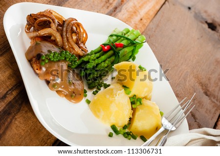 Freshly prepared piece of meat with onions and pepper sauce, cooked potatoes green asparagus and green pepper as a decoration on a white plate.