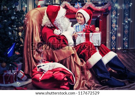 Santa Claus giving a present to a little cute boy near the fireplace and Christmas tree at home.