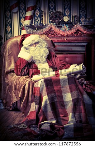 Santa Claus with a list of Christmas presents sitting in a comfortable chair near the fireplace at home.