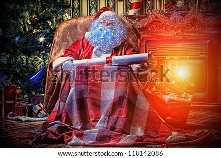 Santa Claus with a list of Christmas presents sitting in a comfortable chair near the fireplace at home.