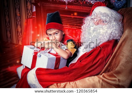 Santa Claus sitting with a little cute boy elf near the fireplace at home.