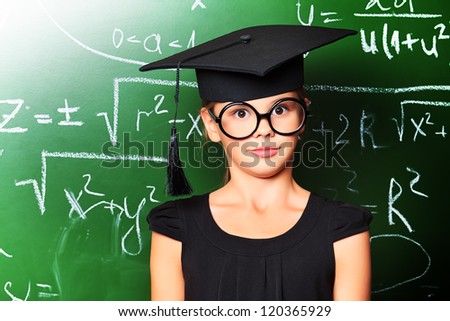 Portrait of a cute schoolgirl in academic hat and big round spectacles at a classroom.