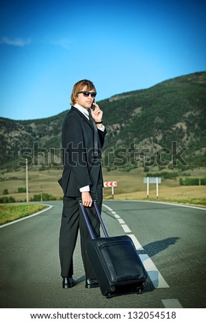 Handsome business man standing on a highway with his suitcase and calling by phone.