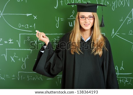 Portrait of a happy graduating student girl in an academic gown standing at the classroom.