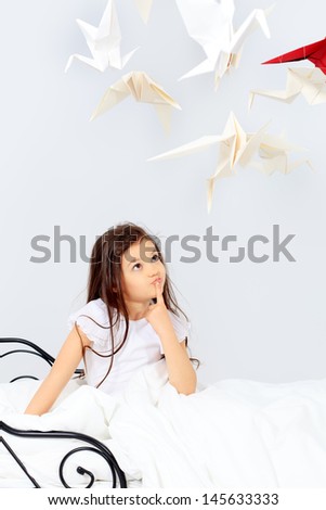 Pretty little girl sitting on her bed surrounded by paper birds. Dream world.