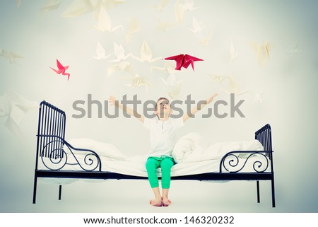 Cute little boy sitting on his bed surrounded by paper birds. Dream world.