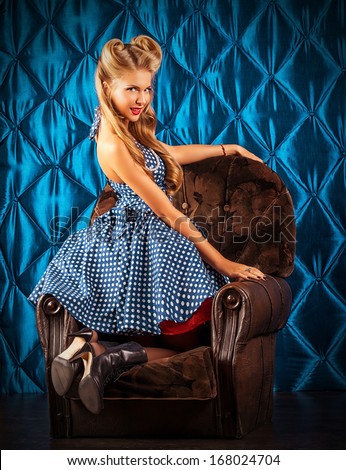 Charming pin-up woman with retro hairstyle and make-up sitting in the armchair over vintage background.