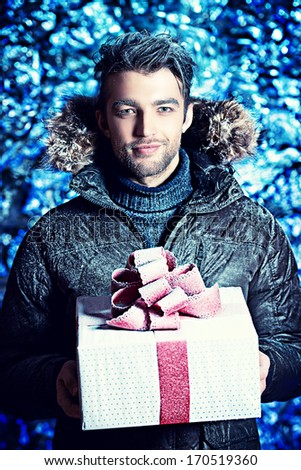 Portrait of a handsome man dressed in winter clothes, holding a gift, covered with snow.