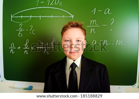 Serious schoolboy in uniform stands near the blackboard at a classroom.
