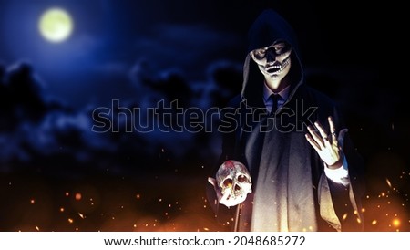 Portrait of a dangerous person with skull makeup dressed in a black robe with a hood holding a human skull in his hands surrounded by fire. Dark moonlit night background with copy space. Halloween. 