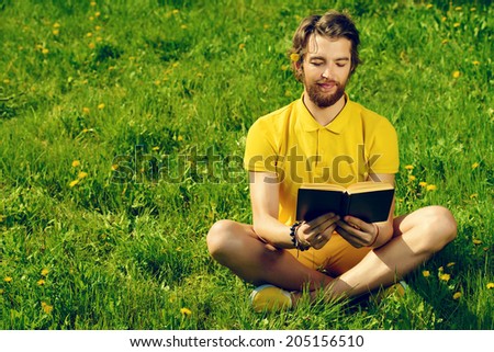 Happy young man sitting on a grass and reading a book. Summer, vacation.
