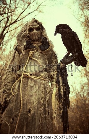 An evil Scarecrow with a canvas bag on his head and in a sackcloth robe stands in a dense forest with a black raven on his hand. Terrible forest sorcerer. Halloween Tales. Horror, thriller.