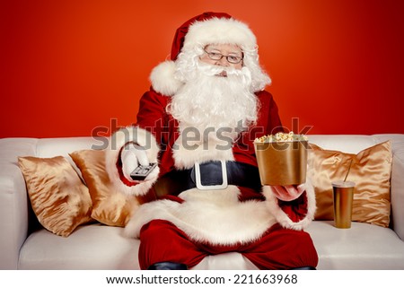 Traditional Santa Claus sitting on the couch watching TV, eating popcorn and drinking soda. Christmas. Red background.