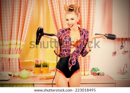 Beautiful pin-up girl doing a hair with a hair dryer and comb on her pink kitchen. Retro style. Fashion.