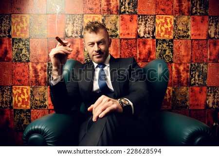 Handsome mature man in elegant suit smoking a cigar. He is sitting on a leather chair in a luxurious interior.