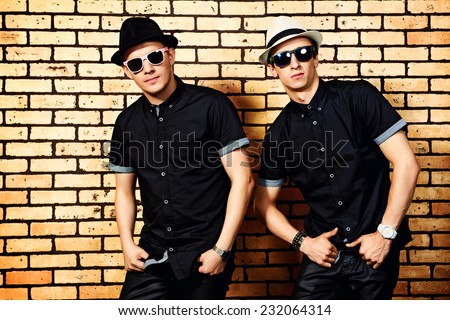 Two handsome men in black shirts and black sunglasses against brick wall.
