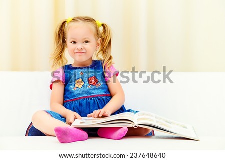 Pretty little girl sitting on a sofa and looking at a children\'s picture book. Happy childhood.