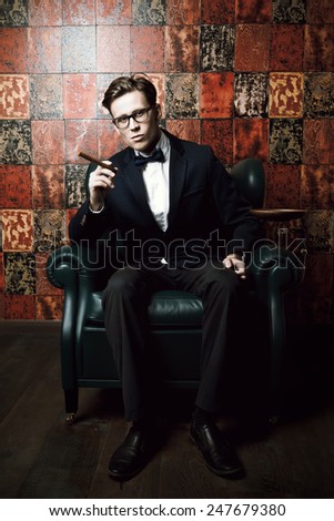 Handsome young man in elegant suit smoking a cigar. He is sitting on a leather chair in a luxurious interior.