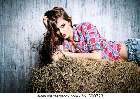 Seductive young woman in jeans shorts and a plaid shirt alluring on a hay. Denim fashion. Western style. Beauty, fashion.