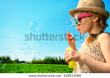 Pretty little girl blows bubbles on a meadow in summer day. Happy childhood. Blue sky.