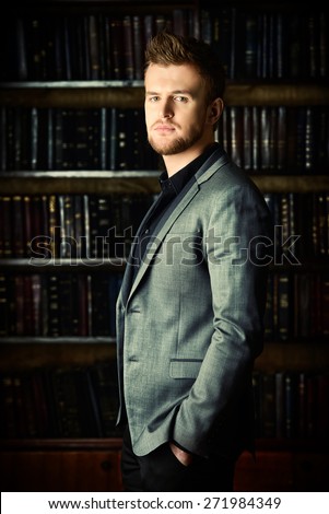 Handsome well-dressed man stands by bookshelves in a room with classic interior. Fashion.