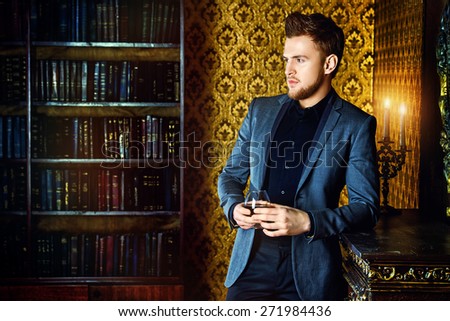 Elegant man in a suit with glass of beverage stands in vintage room. Fashion.