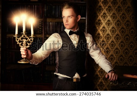 Young handsome man in formal suit and bow-tie stands with candles in a room with classic vintage interior. Fashion. Magic, halloween.