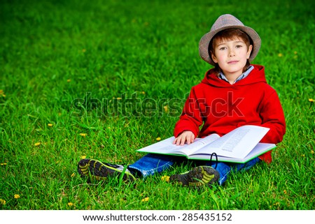 Cute 7 years old boy sitting on a grass with a book. Summer day.