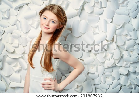 Beautiful blonde teen girl wearing white dress posing by a background of white paper flowers. Beauty, fashion.