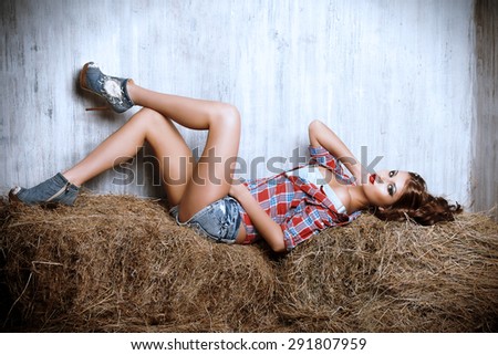 Seductive young woman in jeans shorts and a plaid shirt alluring on a hay. Denim fashion. Western style. Beauty, fashion.