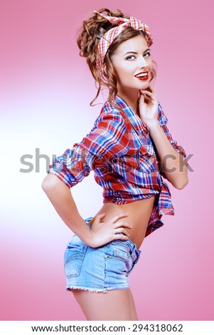 Attractive smiling pin-up girl alluring in shorts and shirt over pink background. Beauty, fashion.