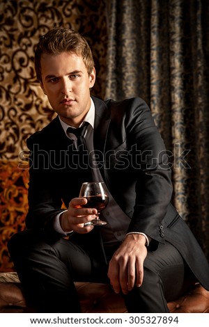 Imposing young man in elegant suit with glass of beverage. Luxury. Vintage interior. Fashion shot.